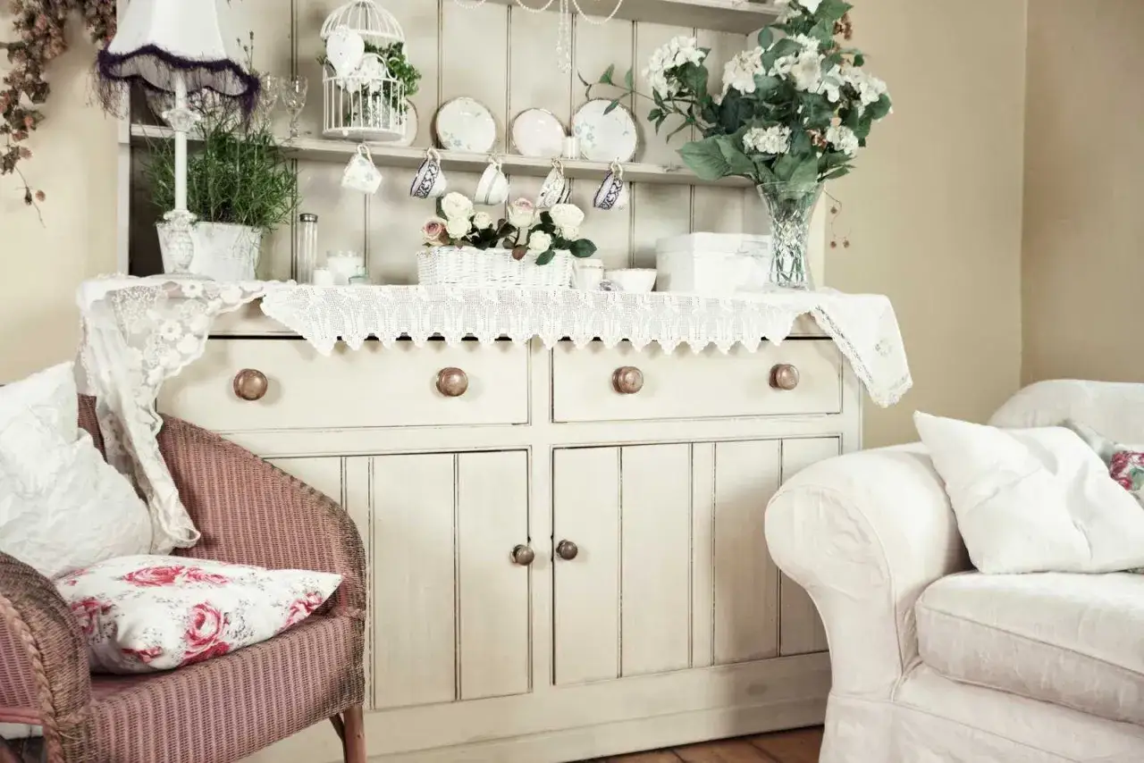 Urocza komoda w stylu shabby chic, ozdobiona koronkami, kwiatami i porcelaną. Obok wiklinowy fotel i biała kanapa.