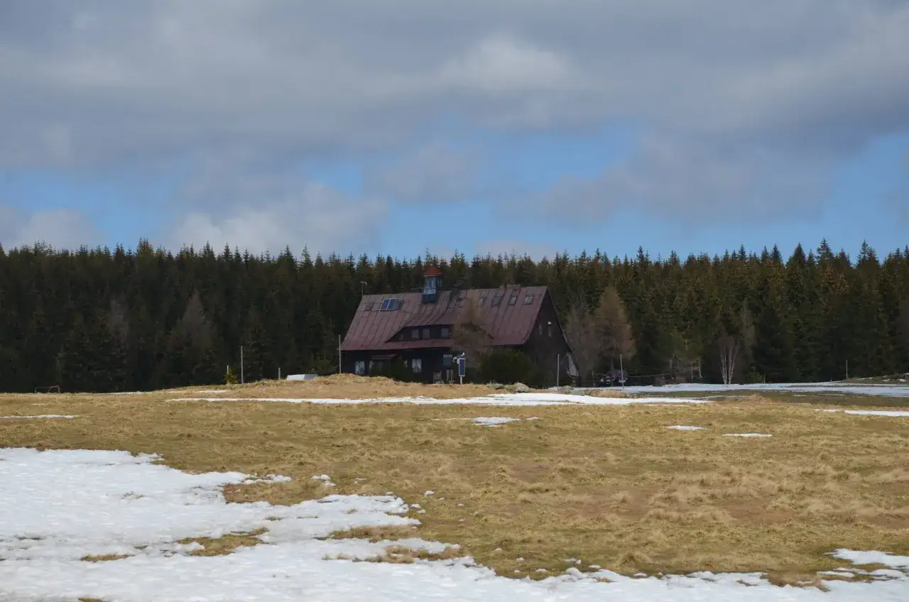 Górzysta chatka z ciemnym dachem, otoczona lasem iglastym. Wiosenna trawa miesza się ze śniegiem.