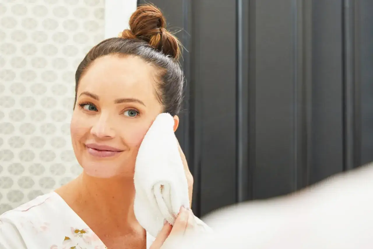 A woman with her hair in a bun gently wipes her face with a soft white towel, enjoying a moment of skin care massage.