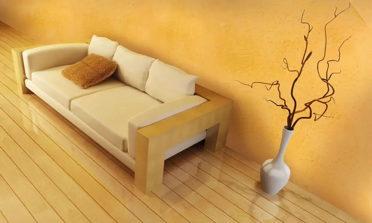 Divano beige con cuscino marrone su parquet lucido, accanto a un vaso bianco con rami secchi. Un'atmosfera che sa di coccio pesto.