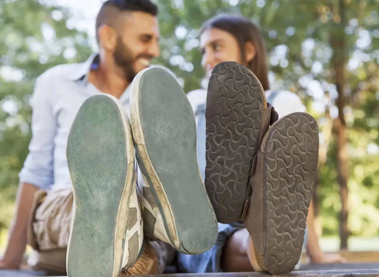 Eva sole, una pareja sonríe mientras sus pies calzados con zapatillas y sandalias descansan al aire libre.
