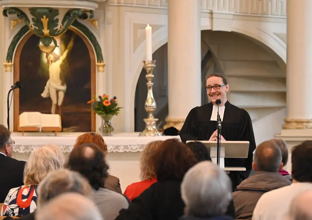 Ein lächelnder evangelischer Pfarrer spricht zu einer Gemeinde in der Kirche. Im Hintergrund ist ein Gemälde von Jesus am Kreuz zu sehen.
