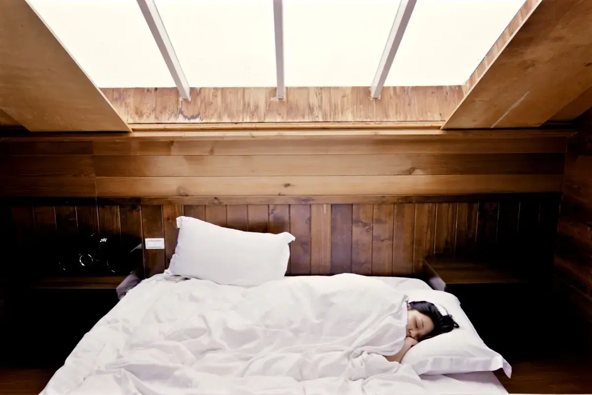Een vrouw slaapt onder witte lakens in een houten kamer. Vraag je je af hoe lang moet een bed luchten?