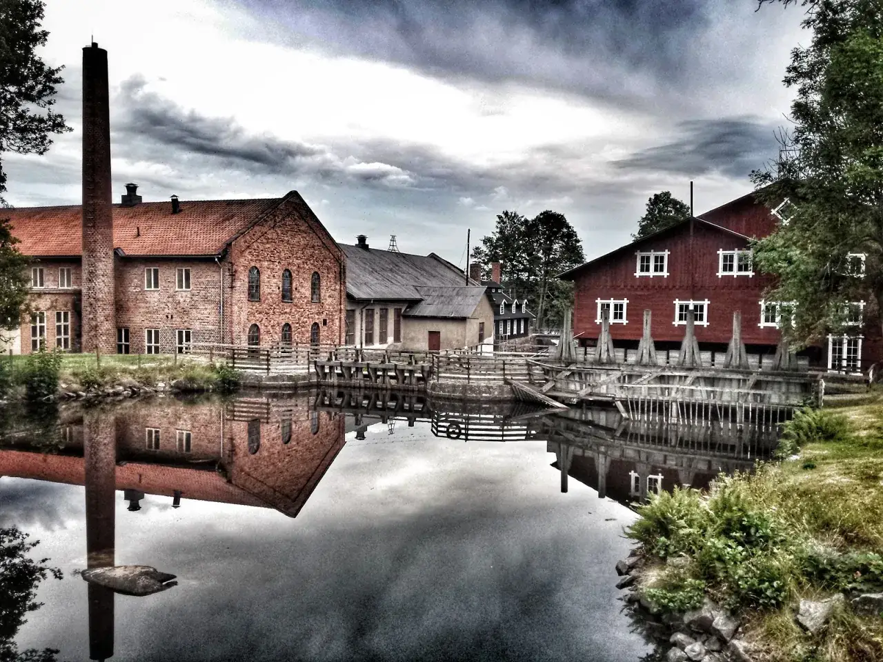 Vattenkvarnar och tegelbyggnader vid forsviks vandrarhem speglas i vattnet under en dramatisk himmel.