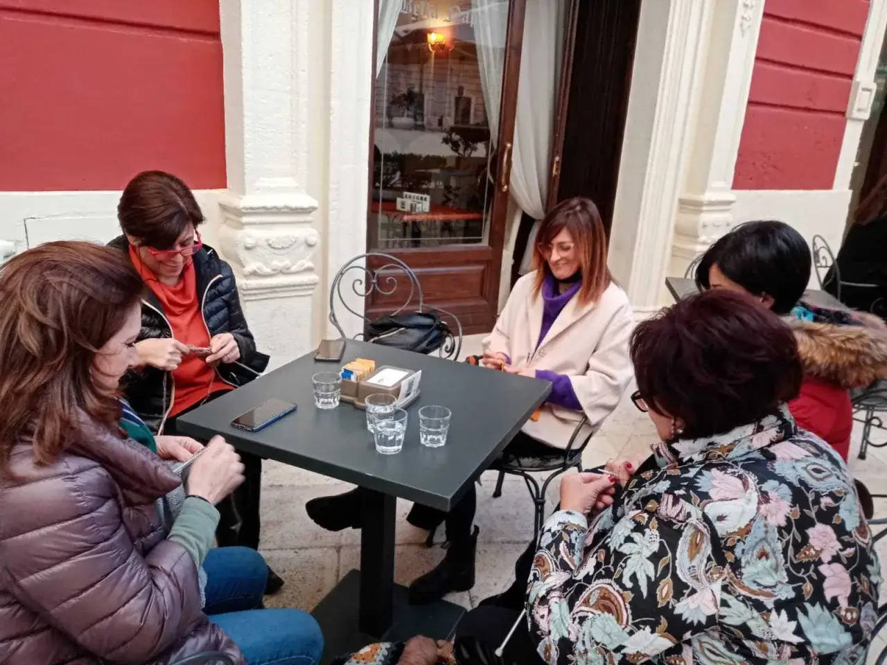 Donne sedute a un tavolo all'aperto, impegnate in un tricot caf&egrave; tutorial. Lavorano a maglia mentre chiacchierano.