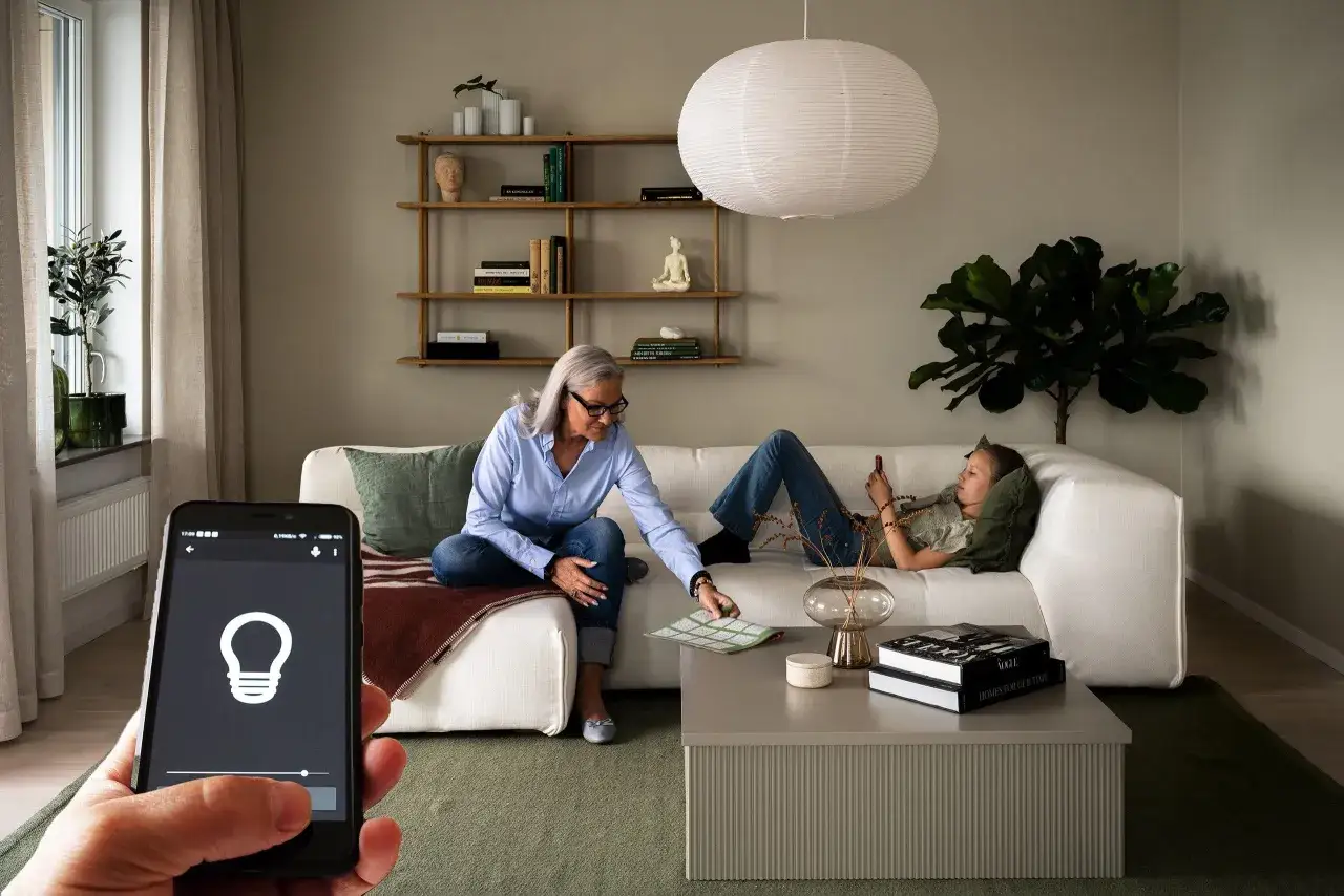 En kvinna och en ton&aring;ring kopplar av i ett modernt vardagsrum. En smartphone visar en app f&ouml;r smart belysning, vilket ger en glimt av hur ser framtiden ut f&ouml;r hemmets bekv&auml;mlighet.