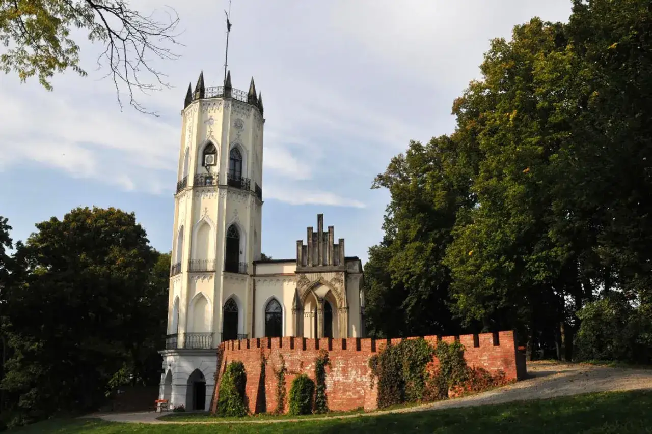 Wieża w stylu neogotyckim, otoczona ceglanym murem i drzewami, przypomina bajkową opinogórę.
