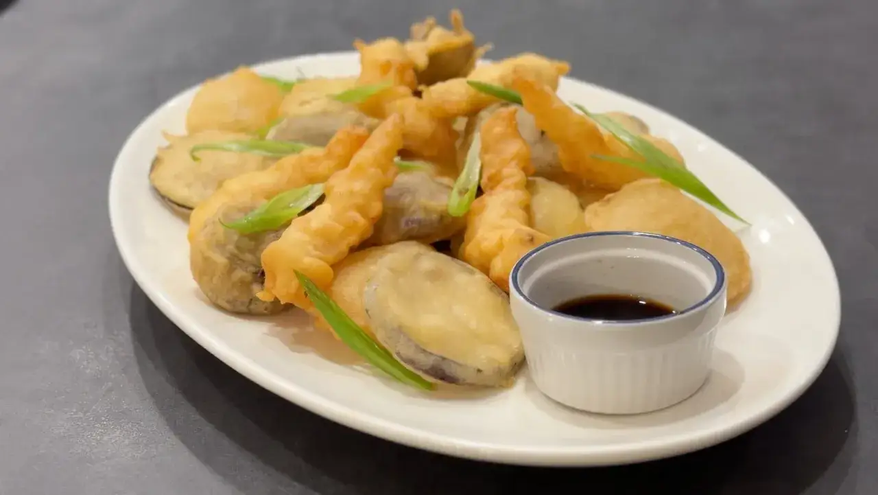 A plate of golden-brown vegetable tempura, featuring slices of eggplant and other fried vegetables, garnished with green onions and served with a small dish of soy sauce.