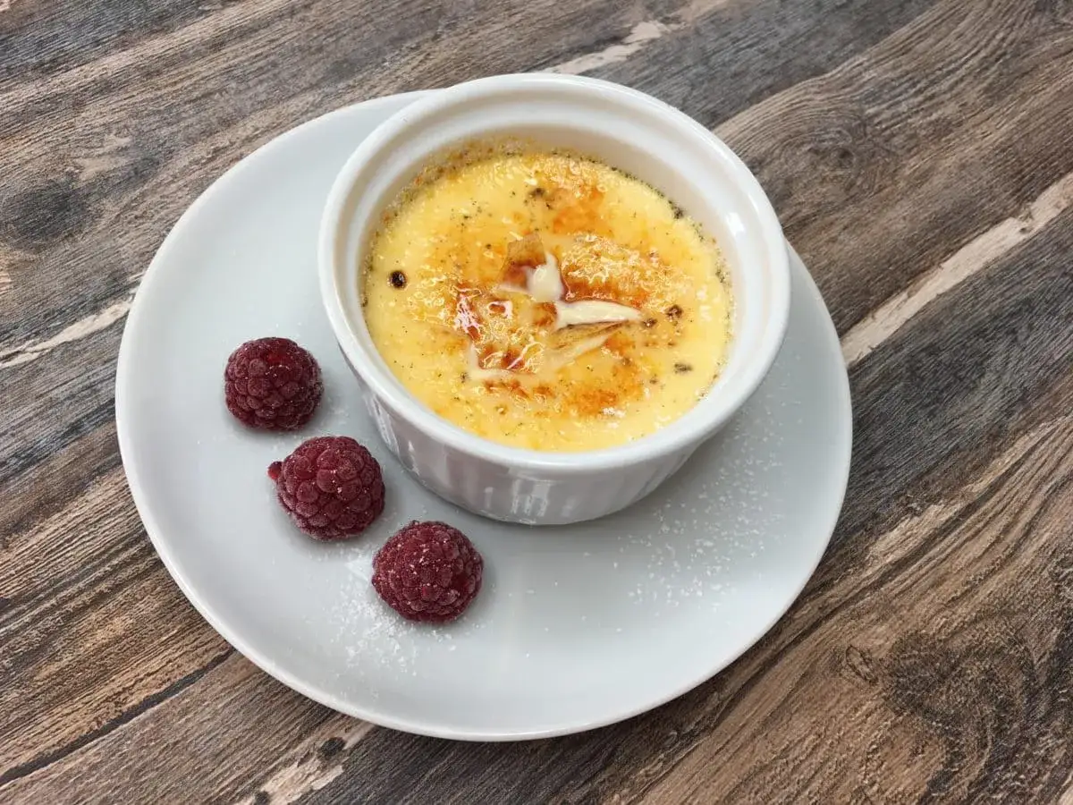 Creme brulee rezept: Ein cremiges Dessert mit karamellisierter Zuckerkruste, garniert mit Himbeeren und Puderzucker auf einem Holztisch.