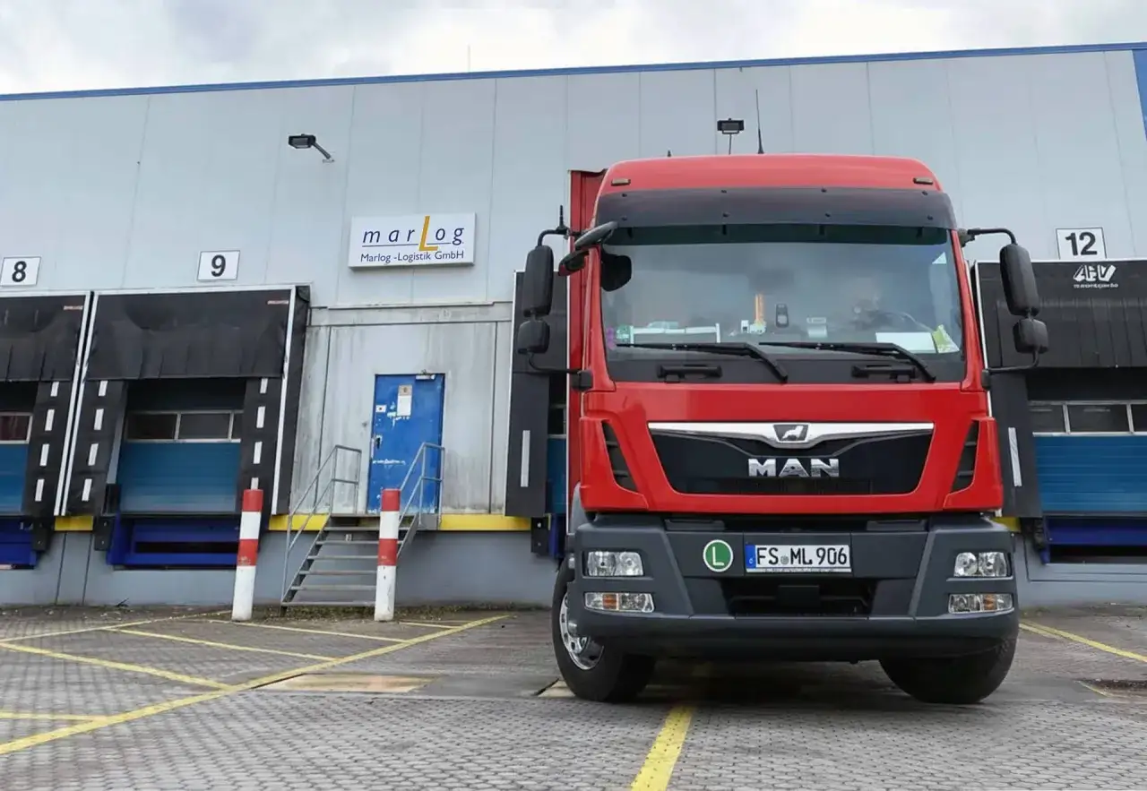 Roter LKW vor einem Lagerhaus mit Laderampen. Die Bedeutung von Distribution wird hier durch die Logistik sichtbar.