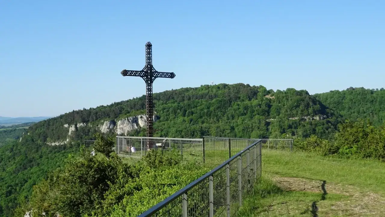 La croix du Dan domine un paysage verdoyant. Une clôture métallique entoure le site, offrant une vue imprenable sur les falaises et la forêt.