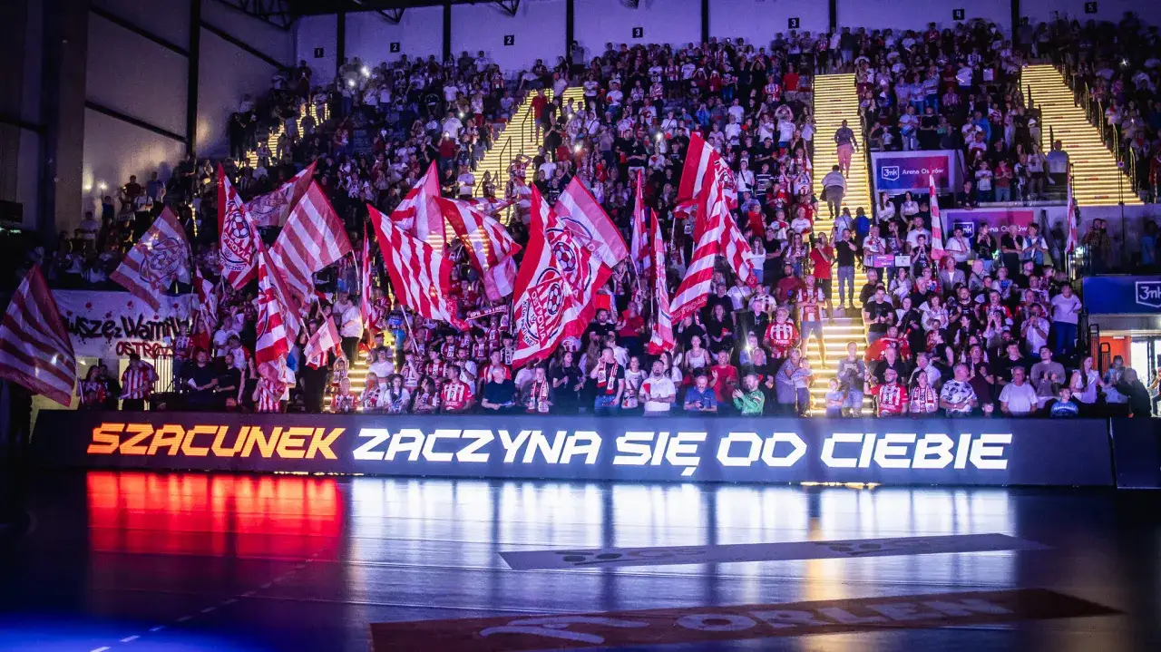 Widownia z flagami kibicuje ostrovia piłka ręczna. Na trybunach tłum w barwach klubowych, na dole napis