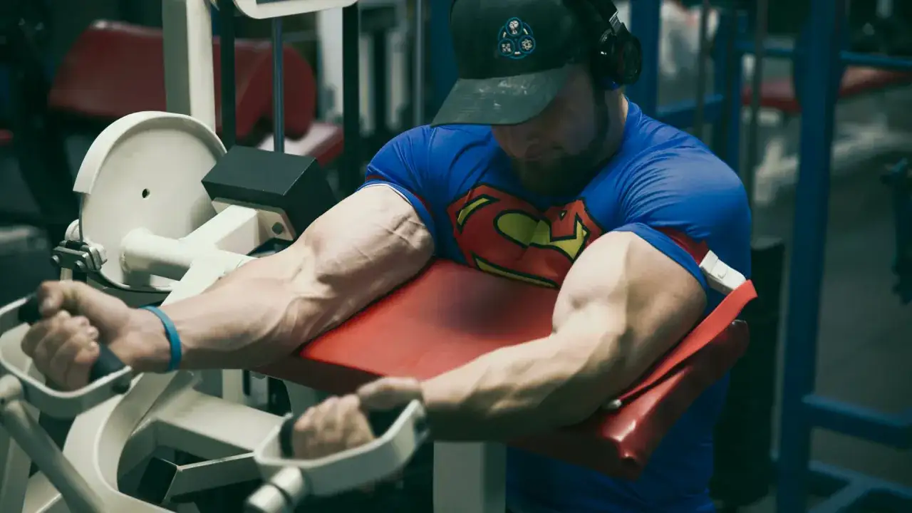 Mięśnie jak u Supermana! Kulturysta w koszulce z logo Supermana ćwiczy bicepsy na maszynie. BFR to trening, który daje takie efekty.