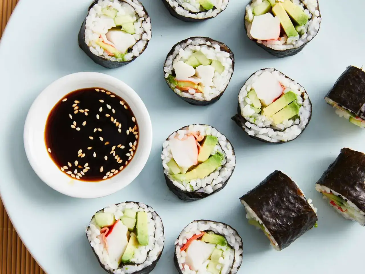 Roladki sushi z awokado, ogórkiem i paluszkami krabowymi, obok miseczka sosu sojowego z sezamem. Idealne składniki do sushi na lekki posiłek.