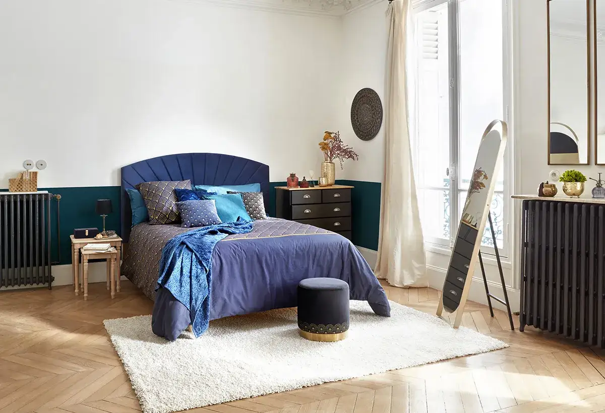 Chambre bleu nuit et dor&eacute;, lit confortable avec coussins assortis, commode noire, miroir sur pied et tapis moelleux.