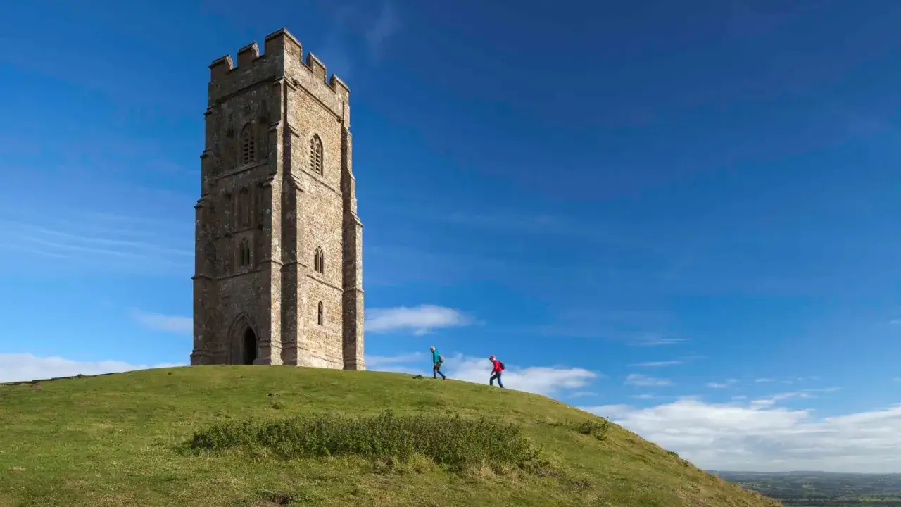 Two hikers ascend the grassy slope towards the iconic Glastonbury Tor tower under a bright blue sky.