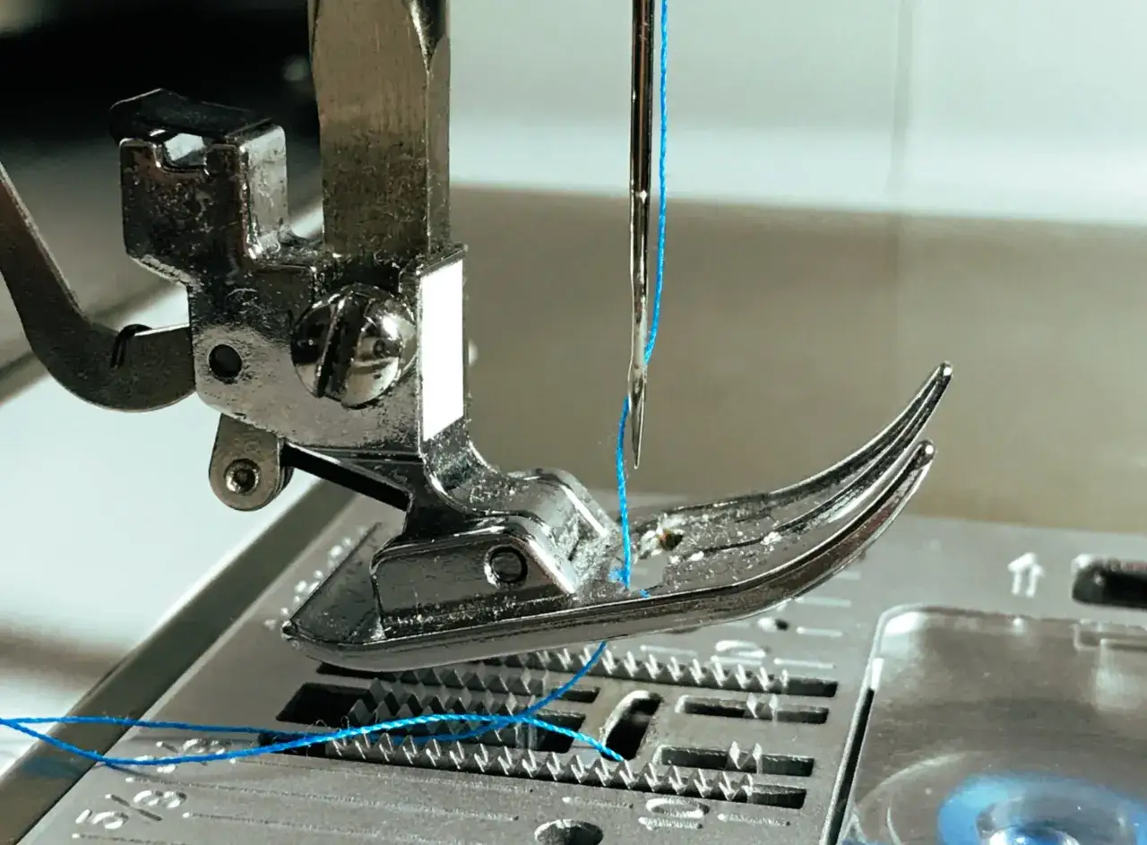 Gros plan sur une aiguille de machine à coudre avec fil bleu, le pied presseur en métal. Un conseil couture pour bien enfiler.
