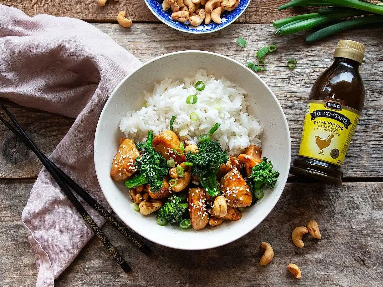 En skål med ris, kyckling och broccoli, toppad med sesamfrön och salladslök. Bredvid syns cashewnötter och en flaska kycklingfond.