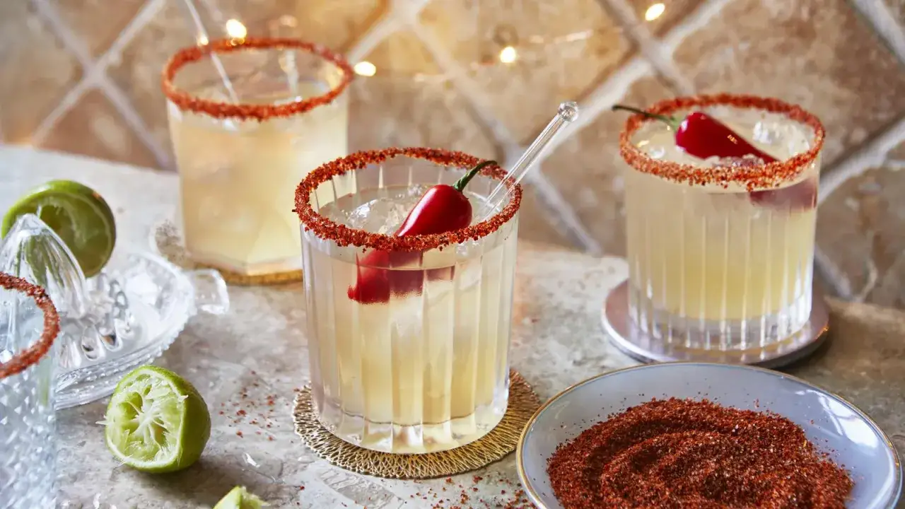Tre glas med en kryddig margarita, dekorerade med chili och saltkant. Lime och en skål med kryddblandning syns också.