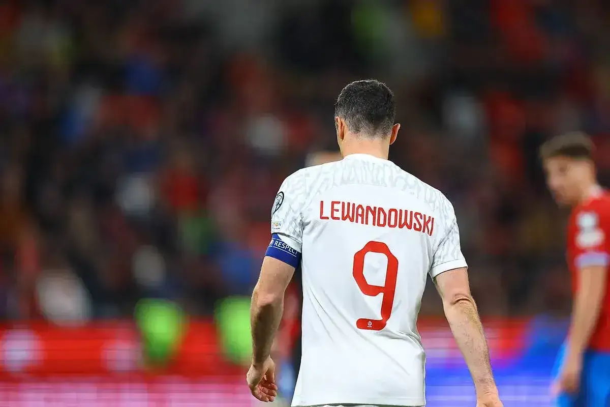 Piłkarze z numerem 9, Robert Lewandowski, w białej koszulce z czerwonym napisem na plecach, na tle trybun.