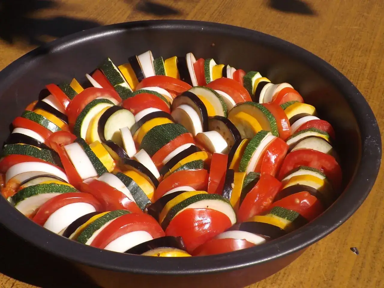 Pr&eacute;paration de la recette ratatouille film : l&eacute;gumes coup&eacute;s en rondelles altern&eacute;es (tomate, courgette, aubergine, oignon) dans un plat.