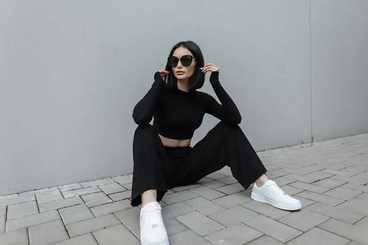 Une femme en tenue noire, crop top et pantalon large, assise sur des pavés. Quelle chaussure avec pantalon large ? Des baskets blanches, parfaites pour ce look décontracté et chic.