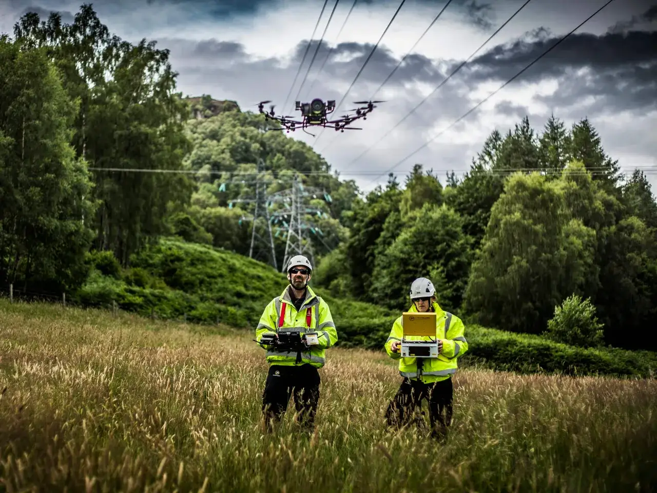 Two technicians in high-visibility jackets operate a power line drone in a grassy field, with a transmission tower in the background.