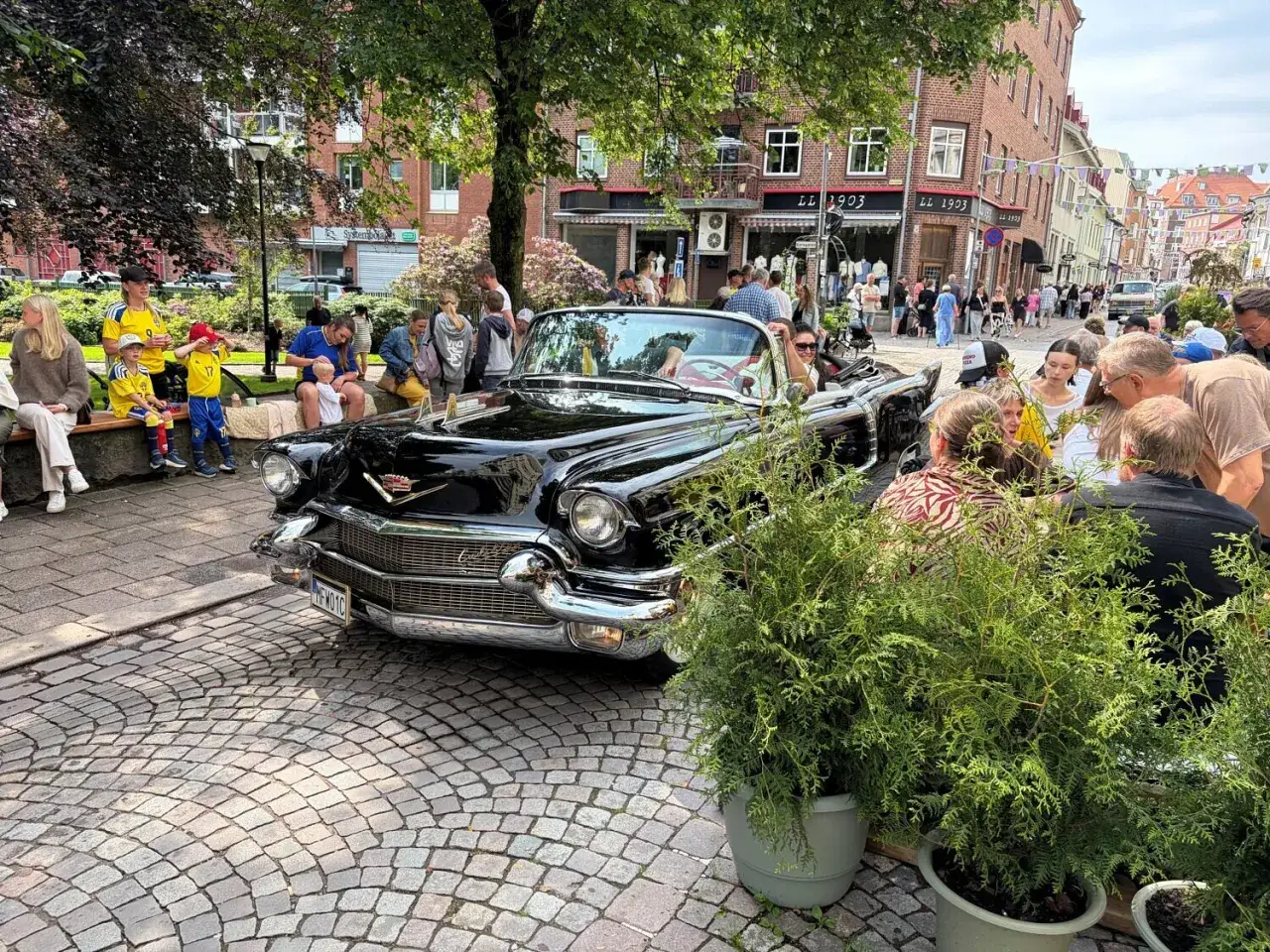 Svart Cadillac cabriolet p&aring; ett kullerstensbelagt torg i Lysekil. M&auml;nniskor samlas runt bilen under ett lyckat lysekil evenemang.