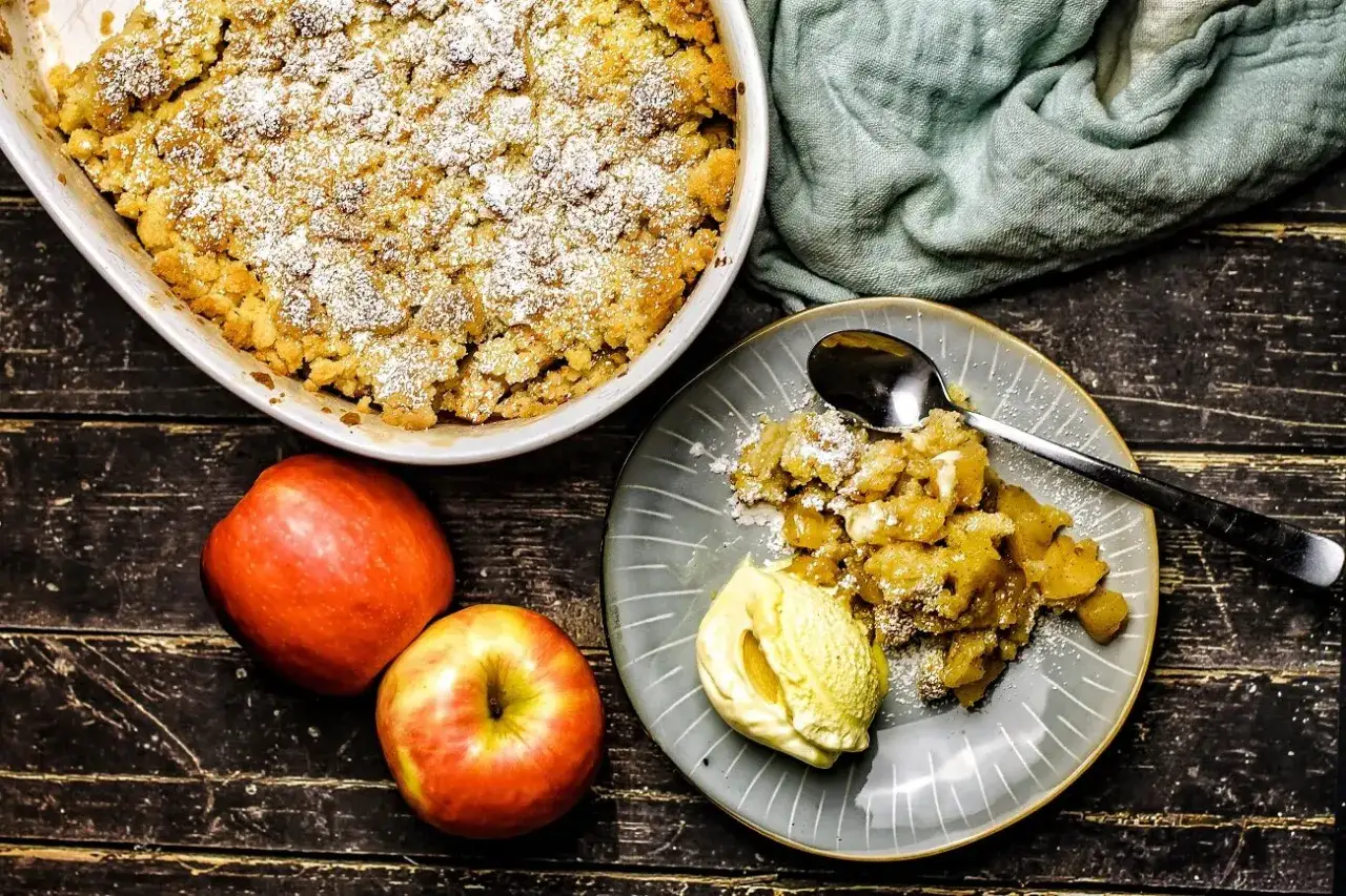 Ein goldbrauner Apfel Crumble, bestäubt mit Puderzucker, serviert mit einer Kugel Vanilleeis. Daneben liegen zwei frische Äpfel.