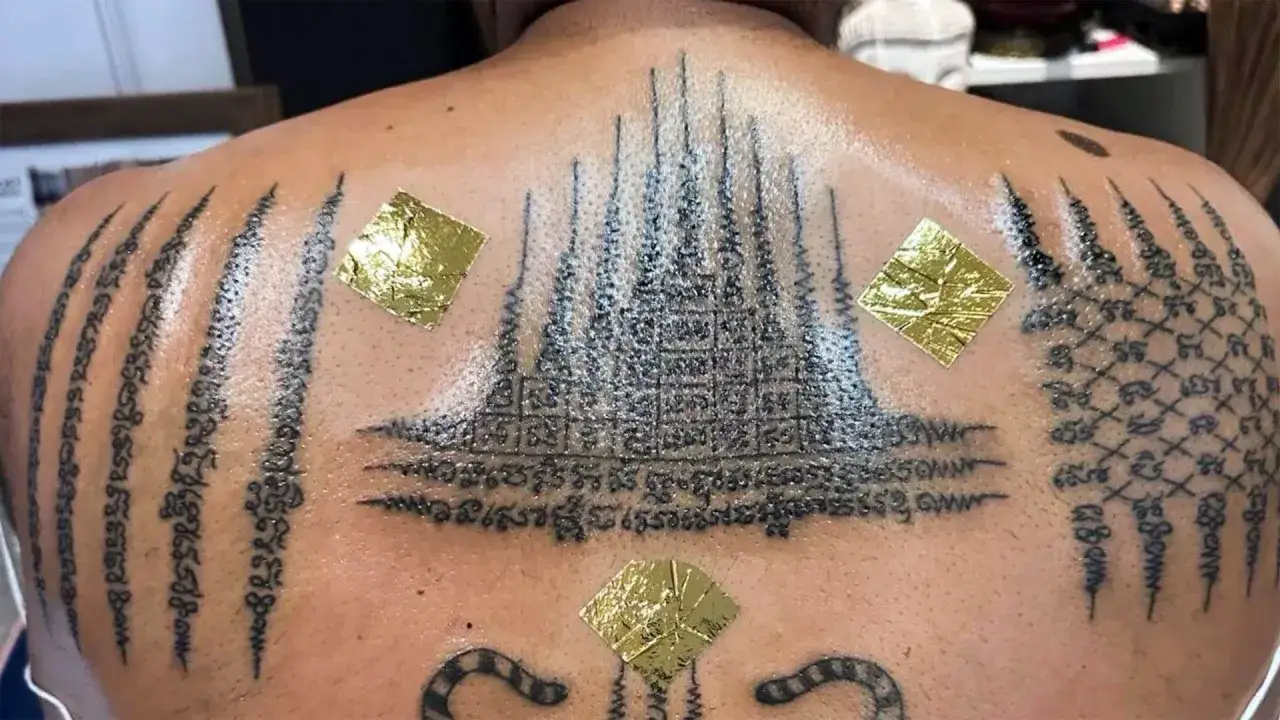 Plecy ozdobione skomplikowanym sak yant tattoo, z geometrycznymi wzorami i buddyjskimi symbolami. Złote płatki dodają mistycznego blasku.