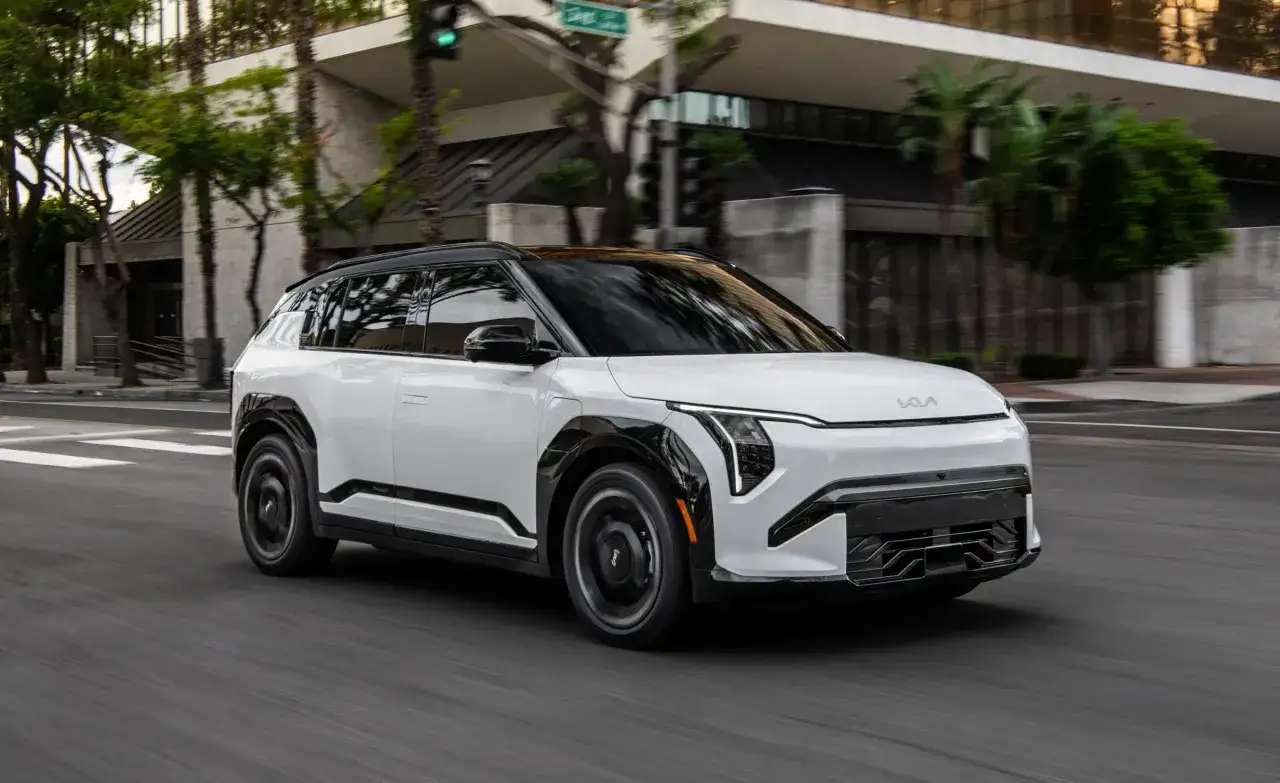 Biały SUV marki Kia, znanej z produkcji amerykańskich samochodów elektrycznych, jedzie ulicą w słoneczny dzień.