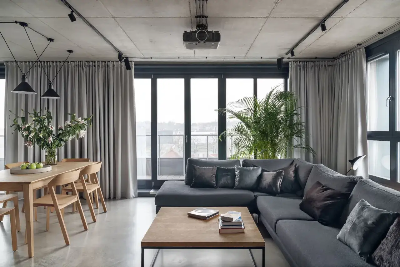 Nowoczesny salon loft inspiracje: szara sofa, drewniany st&oacute;ł, betonowy sufit i duża roślina.
