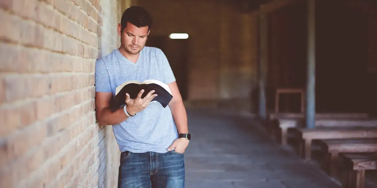Ein Mann lehnt an einer Ziegelwand und liest in seiner evangelischen Bibel.