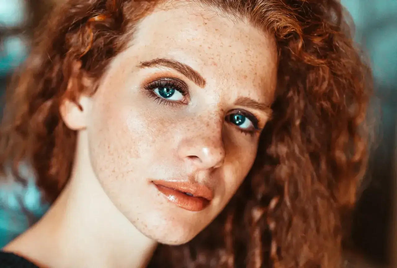 Primo piano di una ragazza con lentiggini e capelli rossi. Il suo sguardo è intenso, con occhi azzurri luminosi. Il trucco accentua il suo viso, con un leggero contouring naso che definisce i lineamenti.