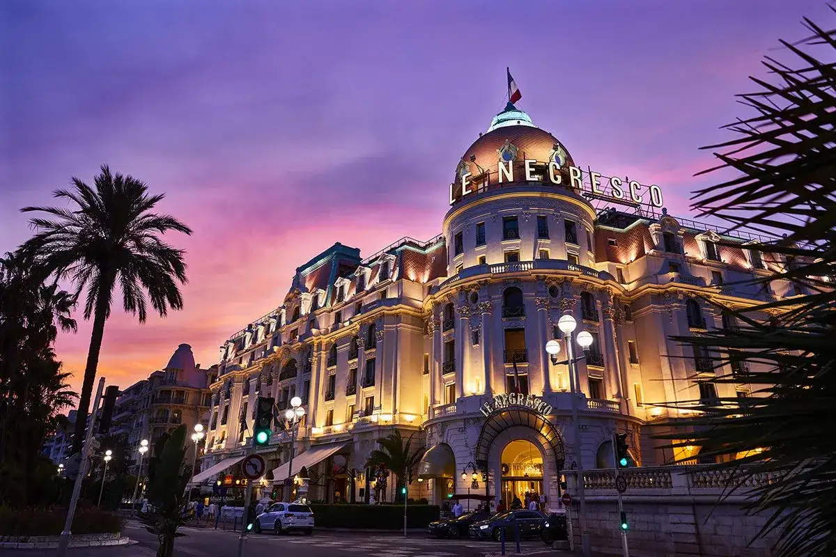 Elegancki hotel Le Negresco w Nicei, oświetlony wieczorem, z palmą i fioletowym niebem.