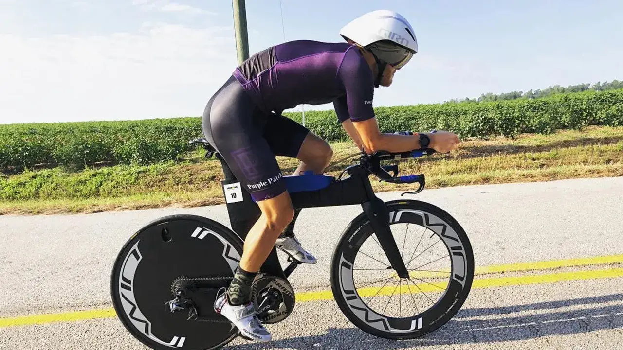 Kolarz w stroju Ironman, w kasku i okularach, ściga się na rowerze aerodynamicznym.