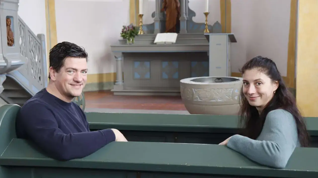 Ein Mann und eine Frau sitzen in einer Kirche. Sie lächeln. Was ist ein Vikar? Ein Vikar ist ein Geistlicher, der einen Pfarrer vertritt.
