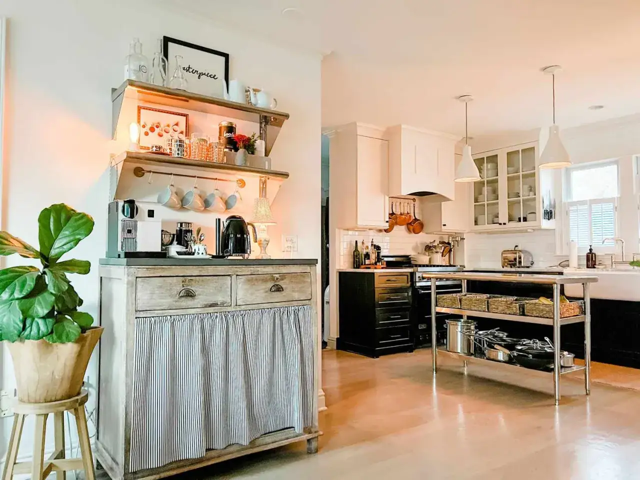 Gemütliche Kaffeebar zu Hause einrichten: Ein weißer Kaffeevollautomat, schwarzer Wasserkocher und Tassen hängen an einem Regal über einer Kommode mit gestreiftem Vorhang.