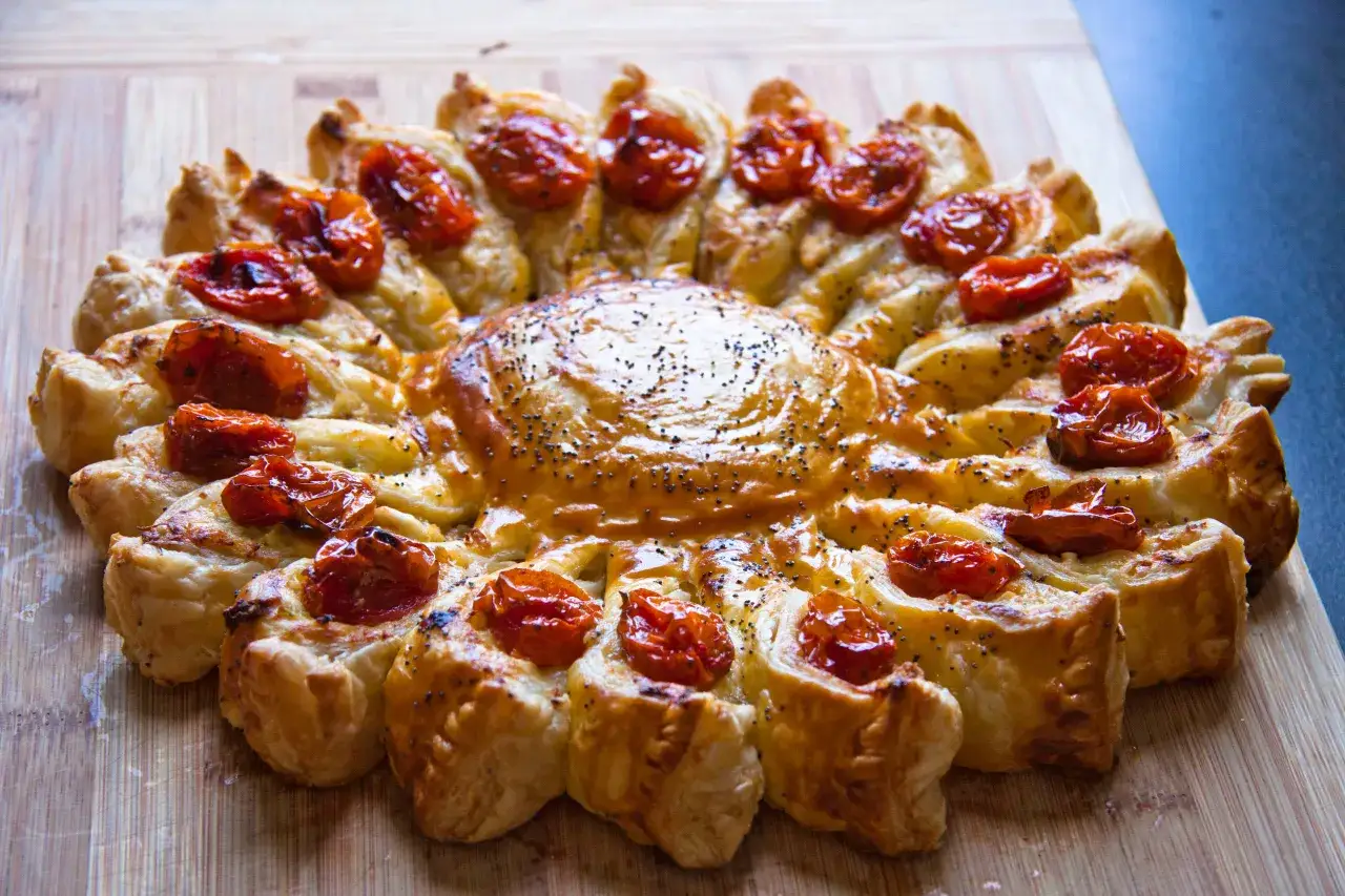 Tarte soleil dor&eacute;e, garnie de tomates cerises confites et de graines de pavot, sur une planche en bois.