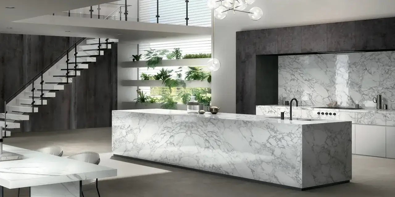 Cucina moderna con isola centrale e rivestimenti in laminam o dekton effetto marmo. Scale a vista e piante verdi creano un ambiente di design.