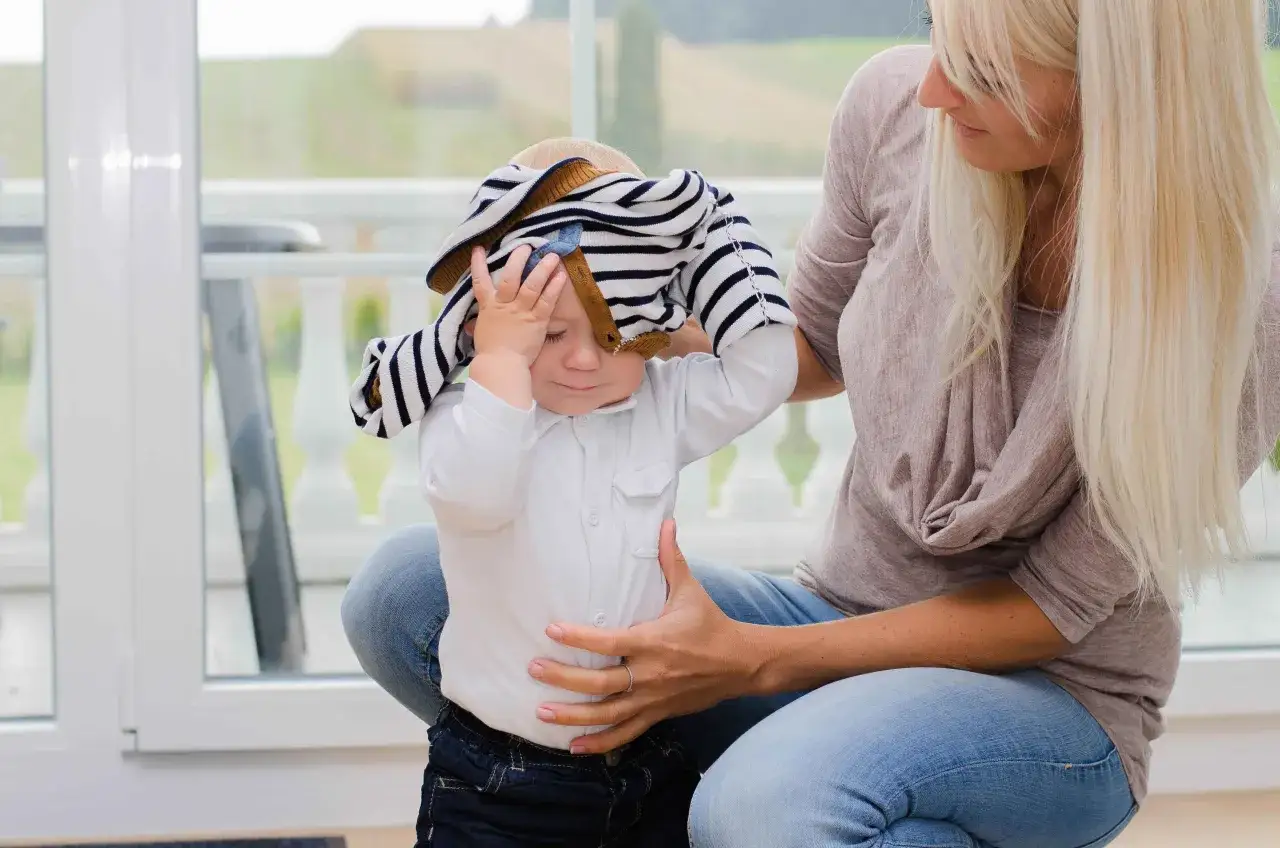 Mutter versucht, ihrem Kind ein gestreiftes Oberteil &uuml;ber den Kopf zu ziehen, aber das **Kind will sich nicht anziehen** und h&auml;lt sich die Stirn.