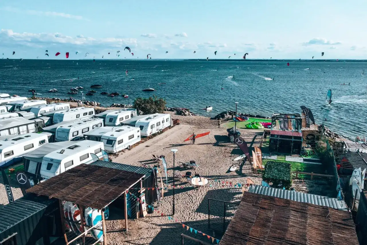 Na plaży pełnej przyczep kempingowych, miłośnicy sportów wodnych szykują się do akcji. W tle widać liczne żagle i latawce na wodzie.
