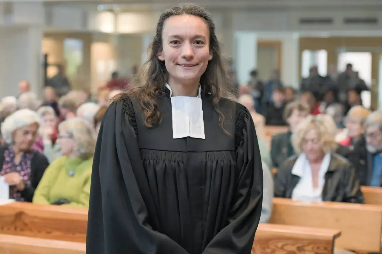 Eine junge evangelische Pfarrerin mit langen Haaren lächelt in die Kamera, gekleidet in ihre Amtskittel. Im Hintergrund sitzen Gemeindemitglieder auf Kirchenbänken.
