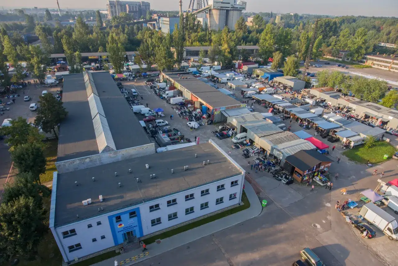 Tłum ludzi i samochody na giełdzie samochodowej. Widać, że giełda samochodowa jest czynna w niedziele, pełna handlarzy i kupujących.