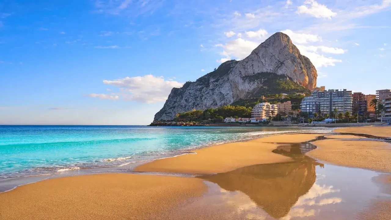 Playa dorada con el imponente Peñón de Ifach al fondo, reflejándose en charcos de agua.
