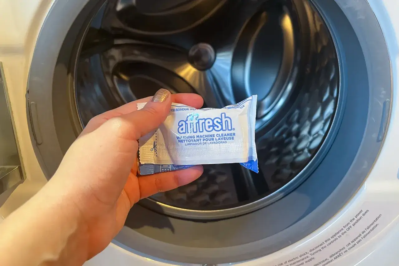 Dłoń trzyma opakowanie środka Affresh, pokazując, jak wyczyścić pralkę. W tle widać wnętrze bębna.