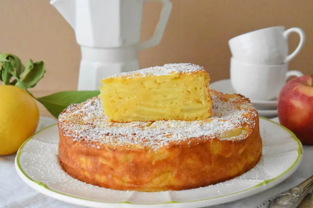 Pyszna torta di mele, posypana cukrem pudrem, z kawałkiem jabłka na wierzchu. Obok cytryna i jabłko.