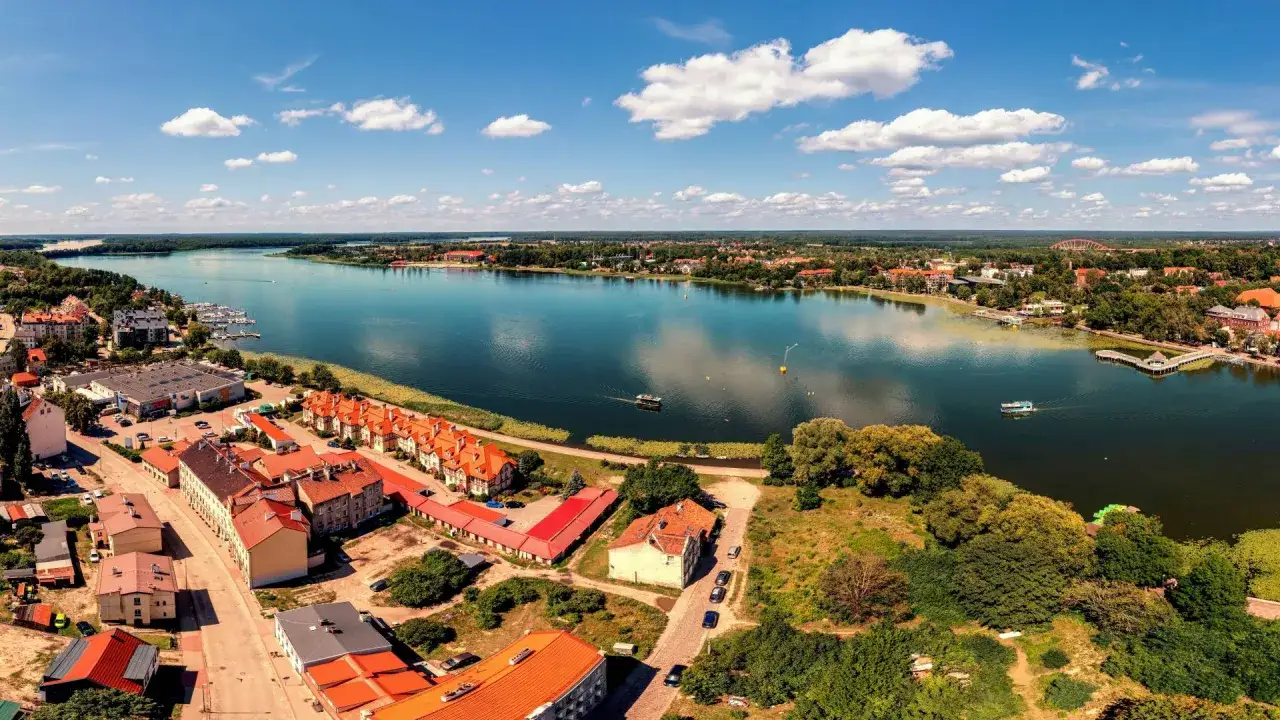 Widok z lotu ptaka na Ostr&oacute;dę i jezioro. Widoczna trasa wzdłuż brzegu, zabudowania i łodzie na wodzie.