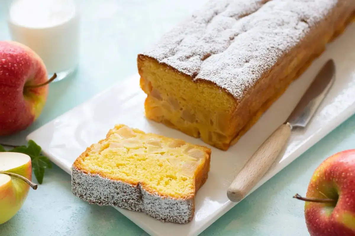 Fetta di plumcake alle mele cosparso di zucchero a velo, accanto a mele rosse e un bicchiere di latte.