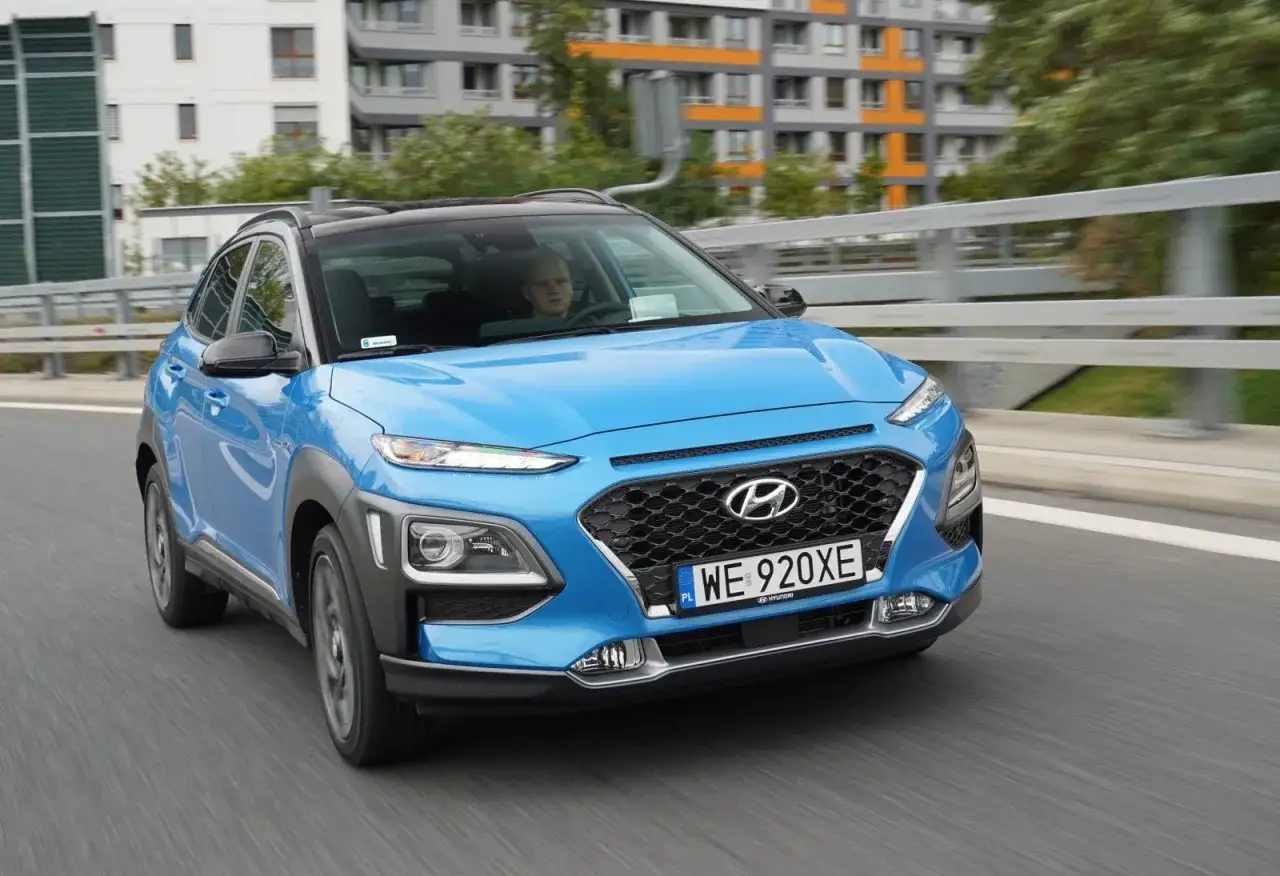 Niebieski Hyundai Kona Hybrid w ruchu miejskim. Kierowca za kierownicą, budynki w tle. Sprawdź jego niskie spalanie!
